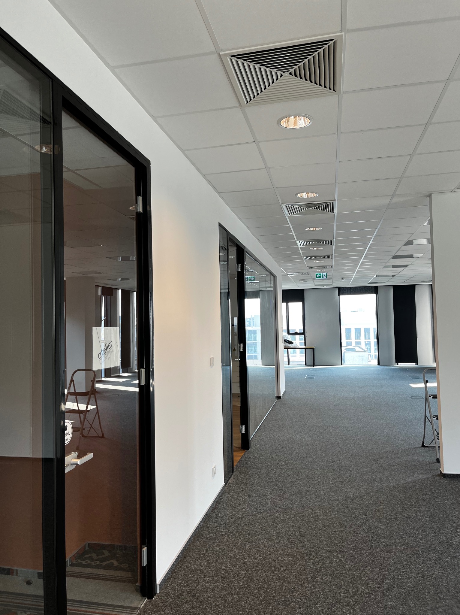 Wnętrze biura z szarą wykładziną, białymi ścianami i czarnymi ramami szklanych drzwi, widok na korytarz z oknami.