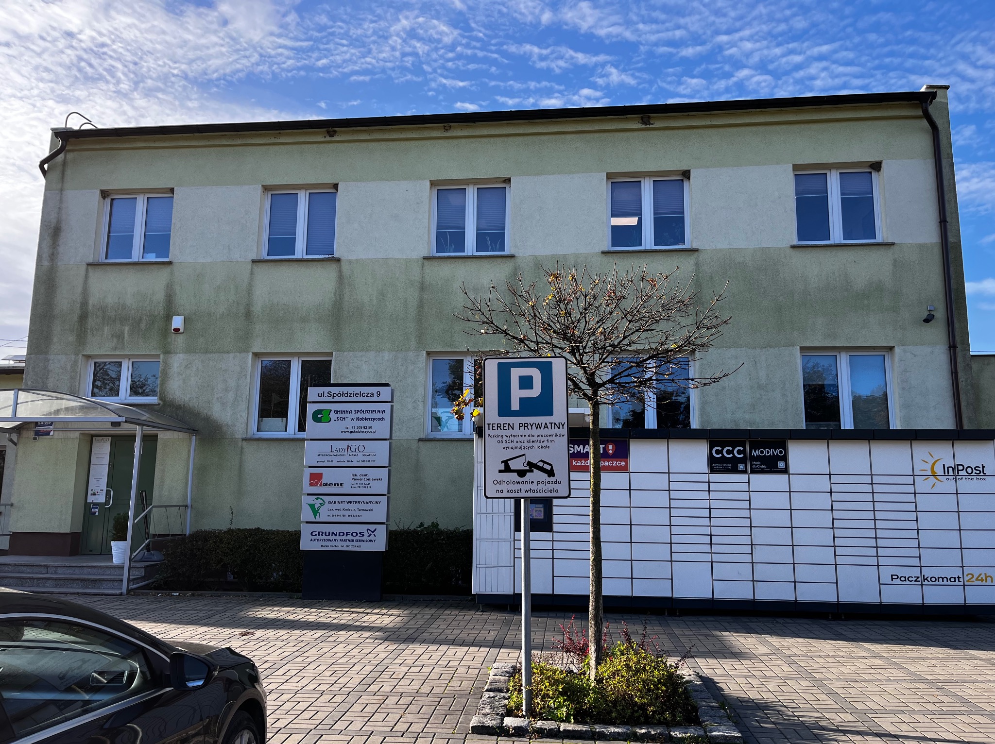Dwupiętrowy budynek biurowy z elewacją w odcieniach beżu i zieleni, z widocznymi oknami, szyldami firm i paczkomatem InPost na pierwszym planie, znak drogowy 'parking' oraz drzewo bez liści.