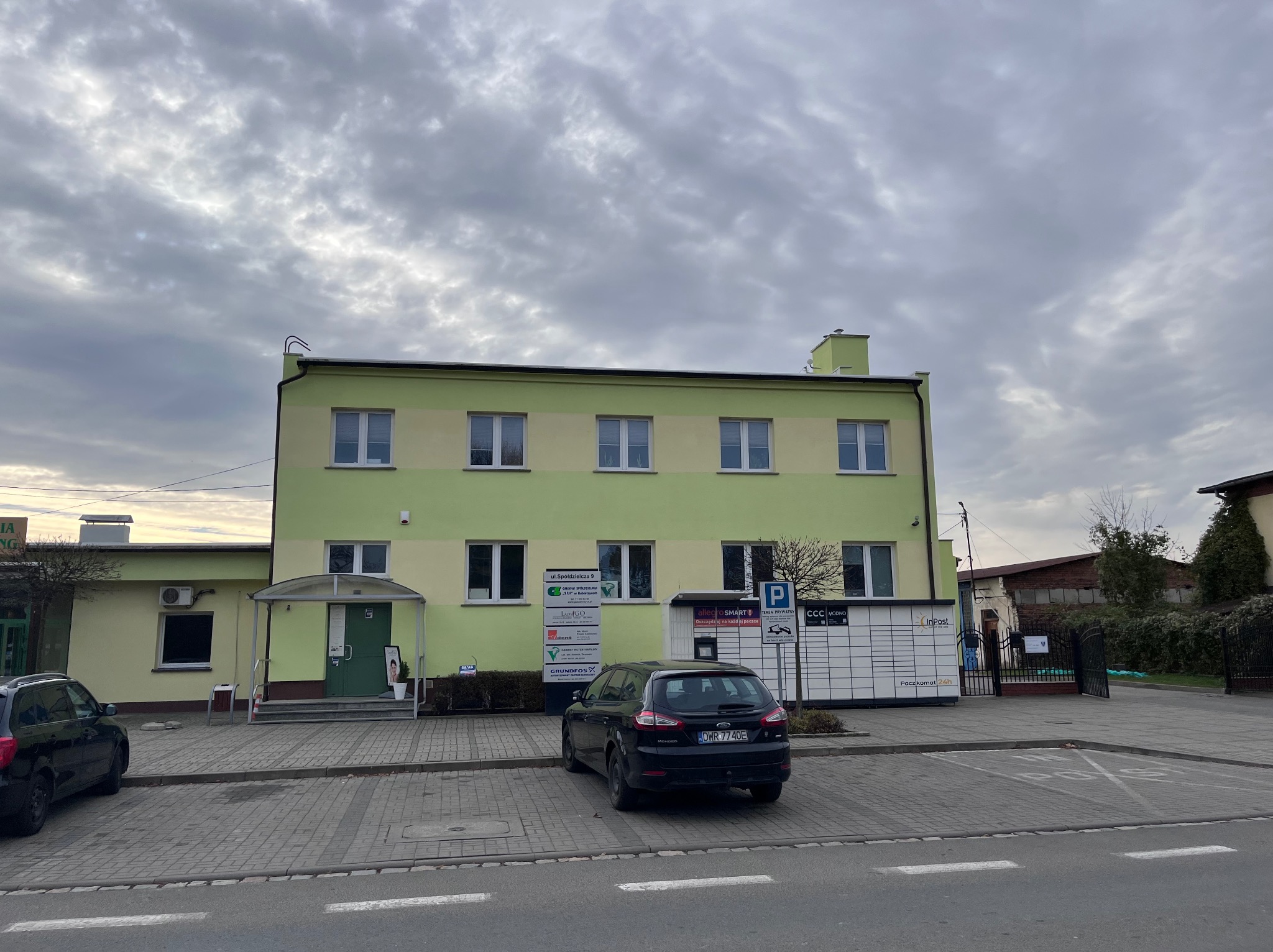 Dwukondygnacyjny budynek biurowy w odcieniach zieleni i beżu, z zaparkowanym czarnym samochodem na pierwszym planie i paczkomatem InPost obok.