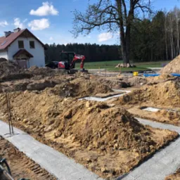 Wylane fundamenty pod dom jednorodzinny z wystającymi prętami zbrojeniowymi, w tle minikoparka i dom w stanie surowym otoczone drzewami.