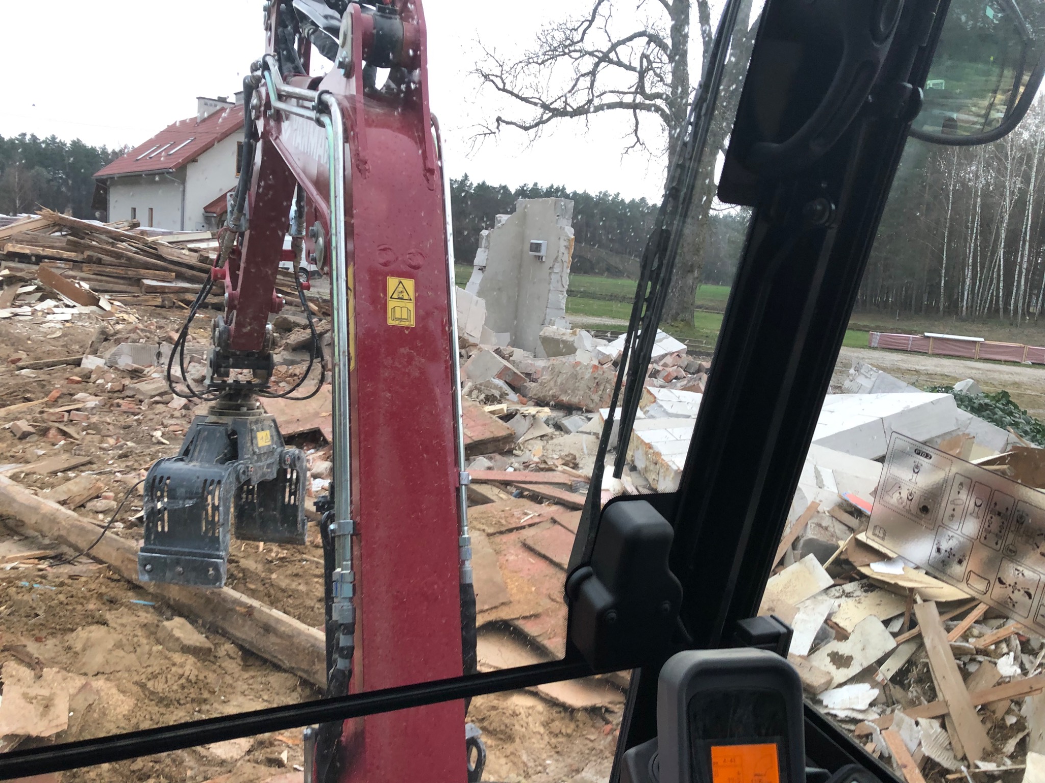 Widok z kabiny czerwonego koparki na teren budowy z gruzem i resztkami budynku, fragment ściany w tle, w oddali dom i drzewa.