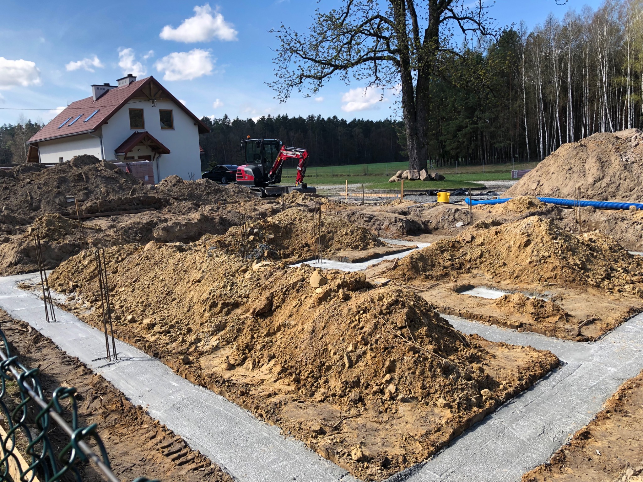 Wylane fundamenty pod dom jednorodzinny z wystającymi prętami zbrojeniowymi, w tle minikoparka i dom w stanie surowym otoczone drzewami.