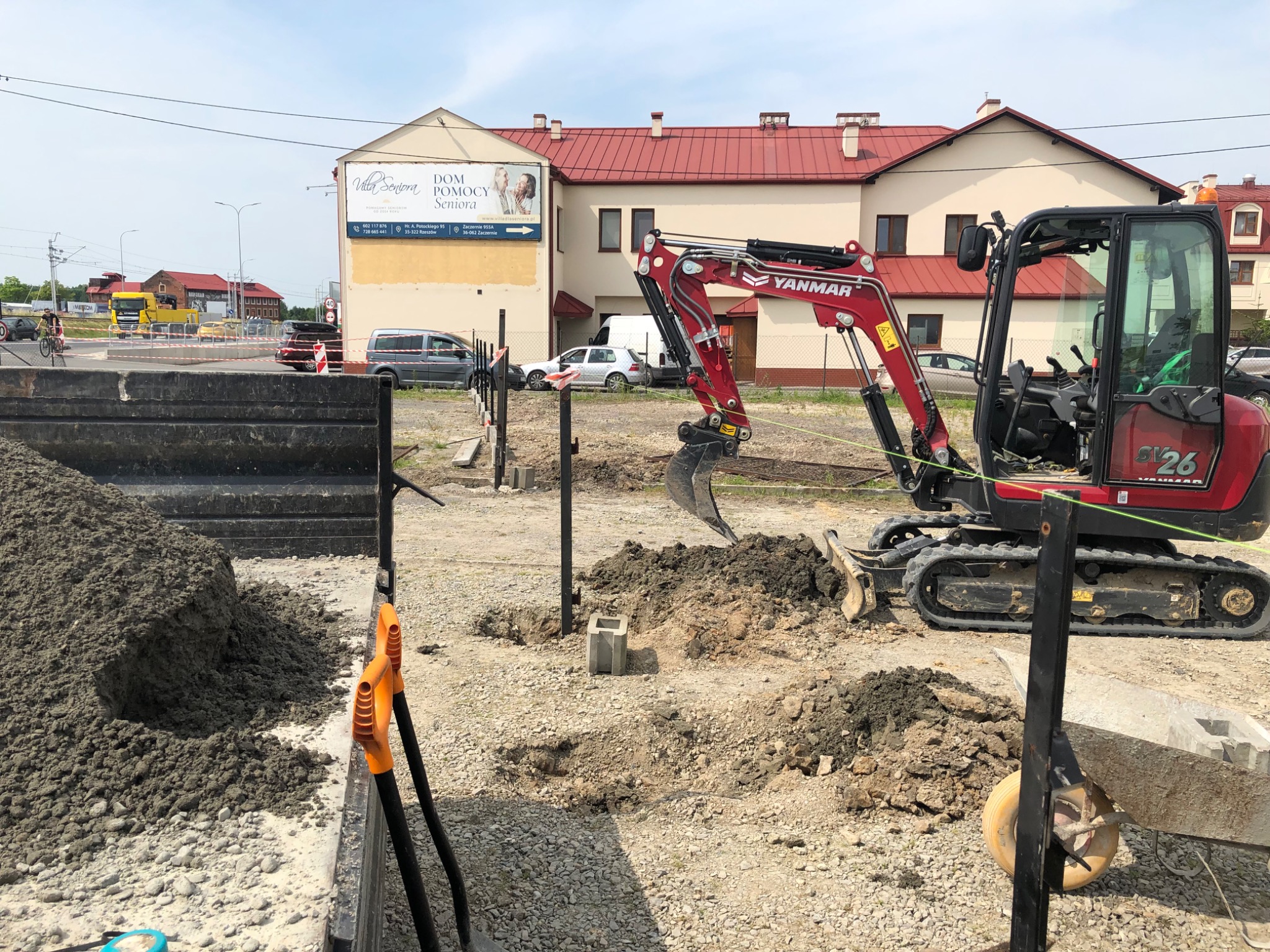 Czerwona mini-koparka Yanmar SL26 w trakcie pracy na placu budowy, widoczne wykopane dołki, stos ziemi i narzędzia, w tle budynek z czerwonym dachem i tablica reklamowa Dom Pomocy Seniora.