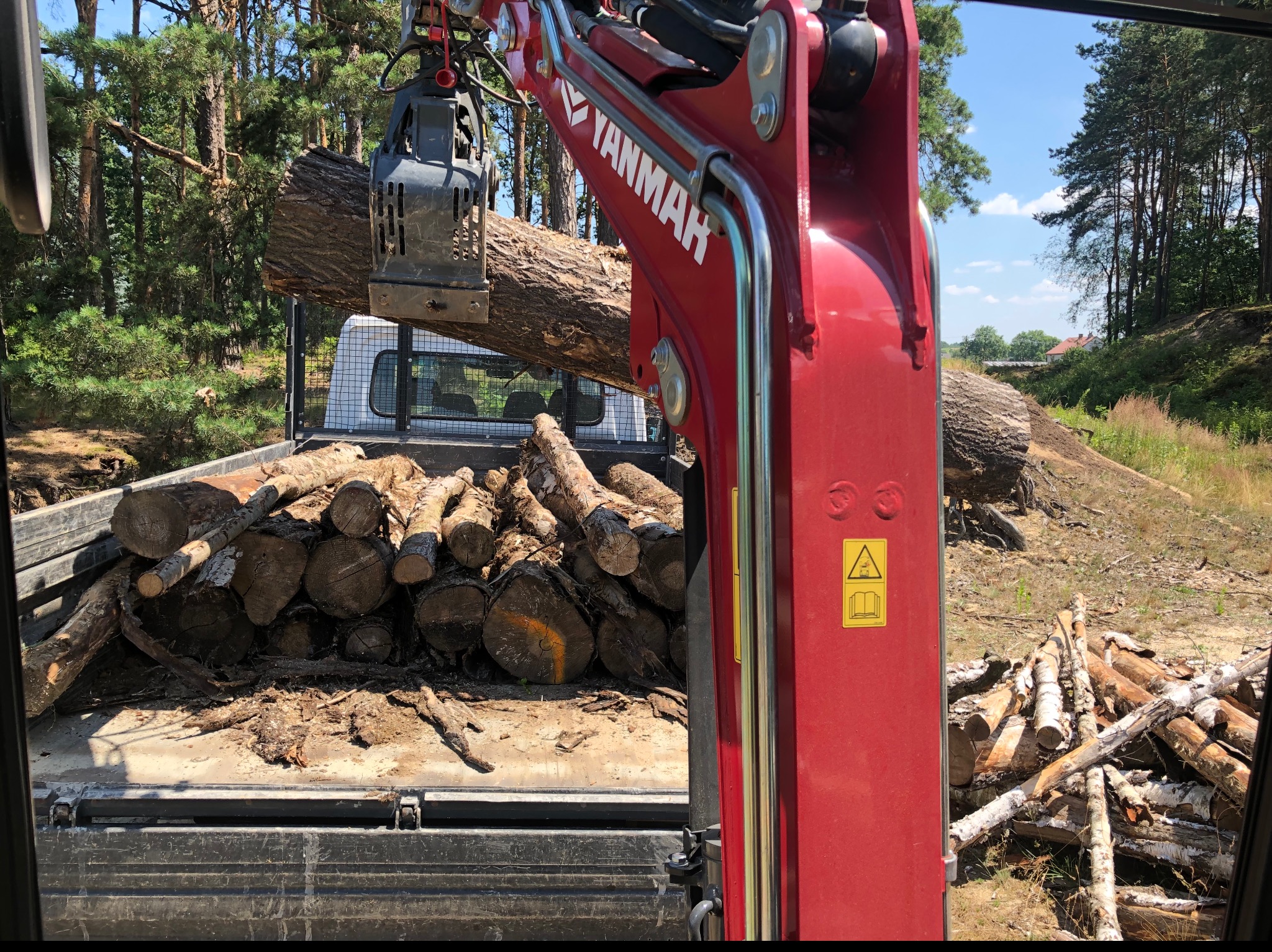 Czerwony minikoparka Yanmar z chwytakiem hydraulicznym podnosi kłodę drewna nad skrzynią załadowaną pociętymi pniami, widok z kabiny na teren zalesiony.