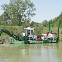 Pływająca koparka, z operatorem w kabinie, pogłębia brzeg stawu, widoczny numer telefonu i adres strony internetowej firmy na burcie.