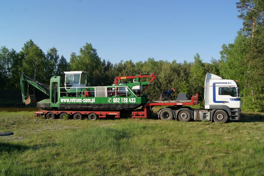 Amfibijna koparka na lawecie transportowana przez ciężarówkę na tle zieleni, z widocznym numerem telefonu i adresem strony internetowej firmy na boku maszyny.