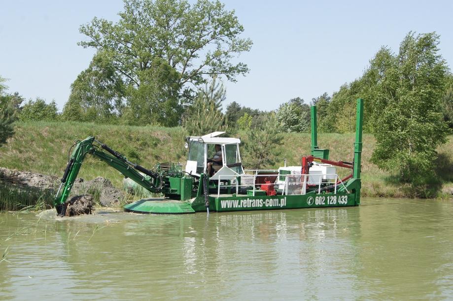 Pływająca koparka, z operatorem w kabinie, pogłębia brzeg stawu, widoczny numer telefonu i adres strony internetowej firmy na burcie.