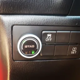 Panel sterowania w samochodzie z przełącznikiem systemu STAG, wskazujący na zasilanie gazem, obok przyciski kontroli trakcji i ustawień ciśnienia w oponach.