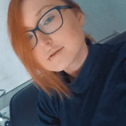 Portret kobiety z rudymi włosami i okularami, ubrana w granatową bluzkę, siedząca na krześle.