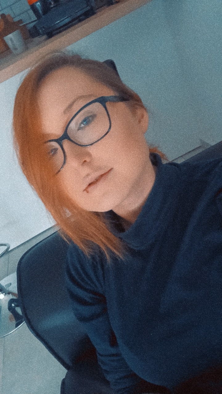 Portret kobiety z rudymi włosami i okularami, ubrana w granatową bluzkę, siedząca na krześle.