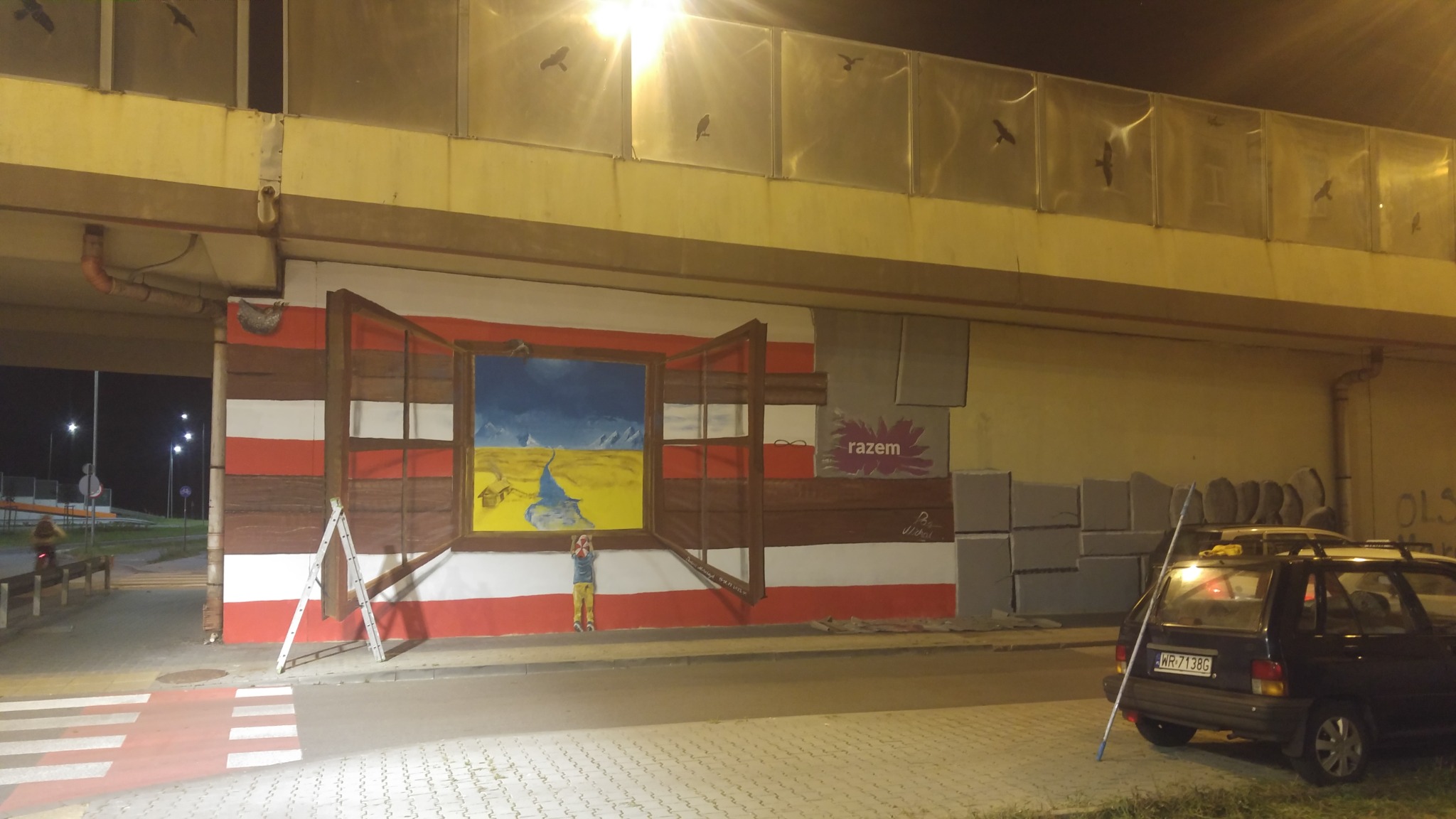 Mural pod wiaduktem przedstawiający otwarte okno z widokiem na krajobraz, z dzieckiem stojącym przed nim i drabiną opartą o ścianę, obok zaparkowany samochód.