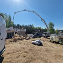 Wylewanie betonu z pompy na budowie domu jednorodzinnego, widoczny szkielet budynku z bloczków, piasek, paleta z workami cementu i samochód transportowy.