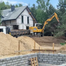Żółta koparka Liebherr nabiera ziemię, obok ciężarówka z wywrotką, w tle dom w stanie surowym otwartym z szarą dachówką, na pierwszym planie fundament z bloczków betonowych i piasek.