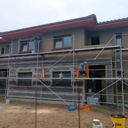 ZAW-TECH Kamil Zawadzki - Dom w trakcie ocieplania, widoczne rusztowania i pracownicy. Część elewacji otynkowana na szaro, okna z brązowymi ramami. Na ziemi wiadra z farbą.