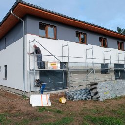 ZAW-TECH Kamil Zawadzki - Dwóch pracowników na rusztowaniu ociepla białą elewację domu z szarą górną częścią. Widoczny styropian, narzędzia i elementy rusztowania. Dom z brązowymi oknami.