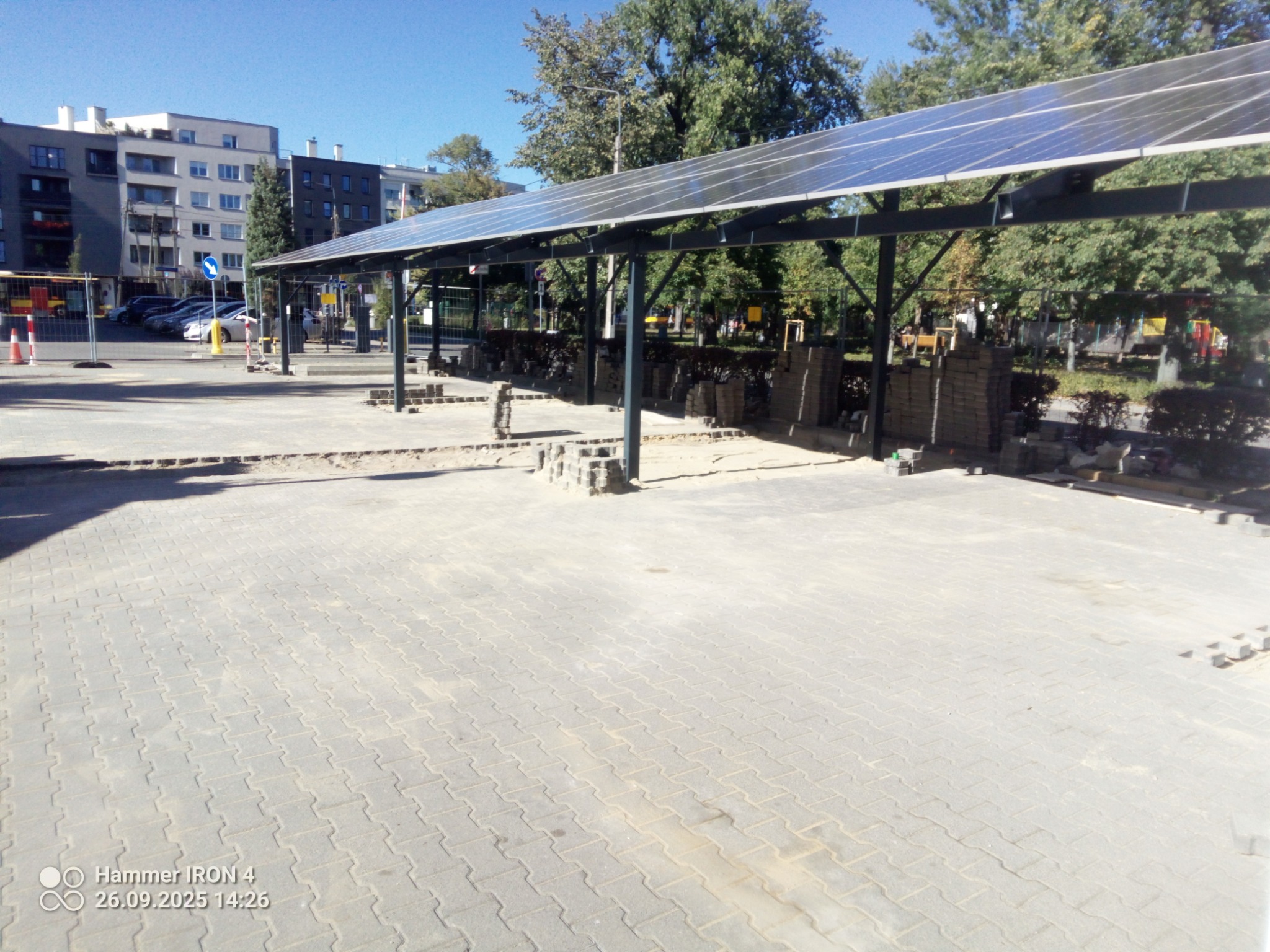Rozległy plac z kostki brukowej w trakcie budowy z zadaszeniem solarnym. Ułożona kostka, palety z materiałem i słupki wskazują na trwające prace.
