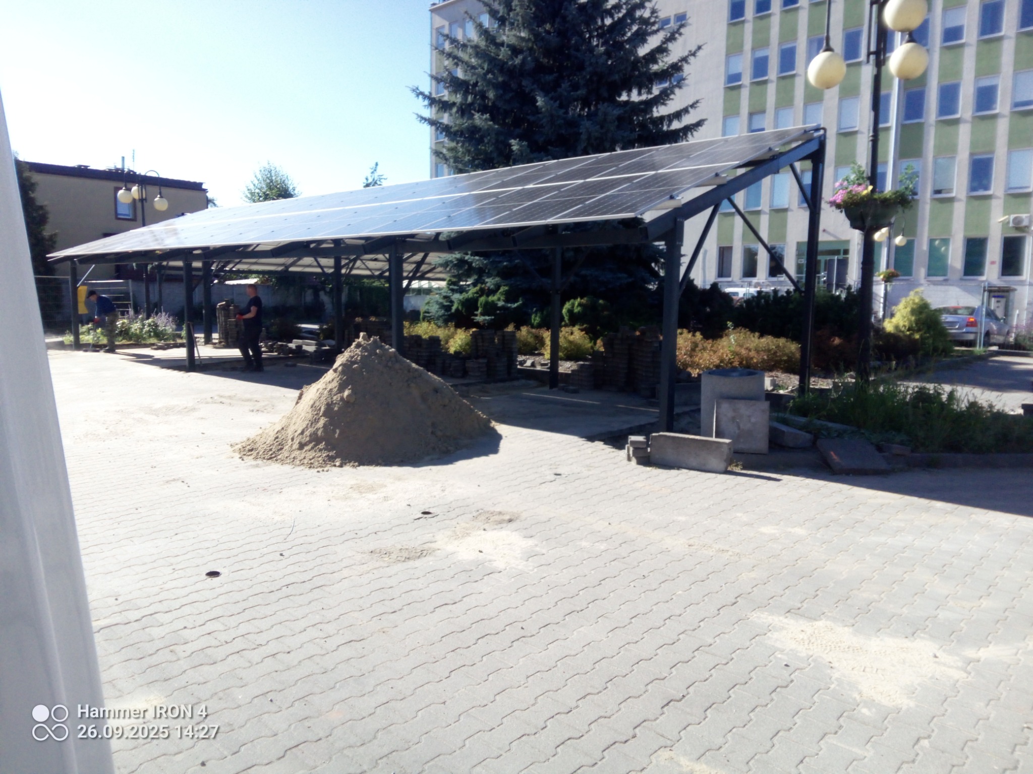 Plac z kostki brukowej w trakcie budowy z zadaszeniem z paneli słonecznych. W tle budynek, drzewa i pracownicy. Widoczna sterta piasku i ułożona kostka.