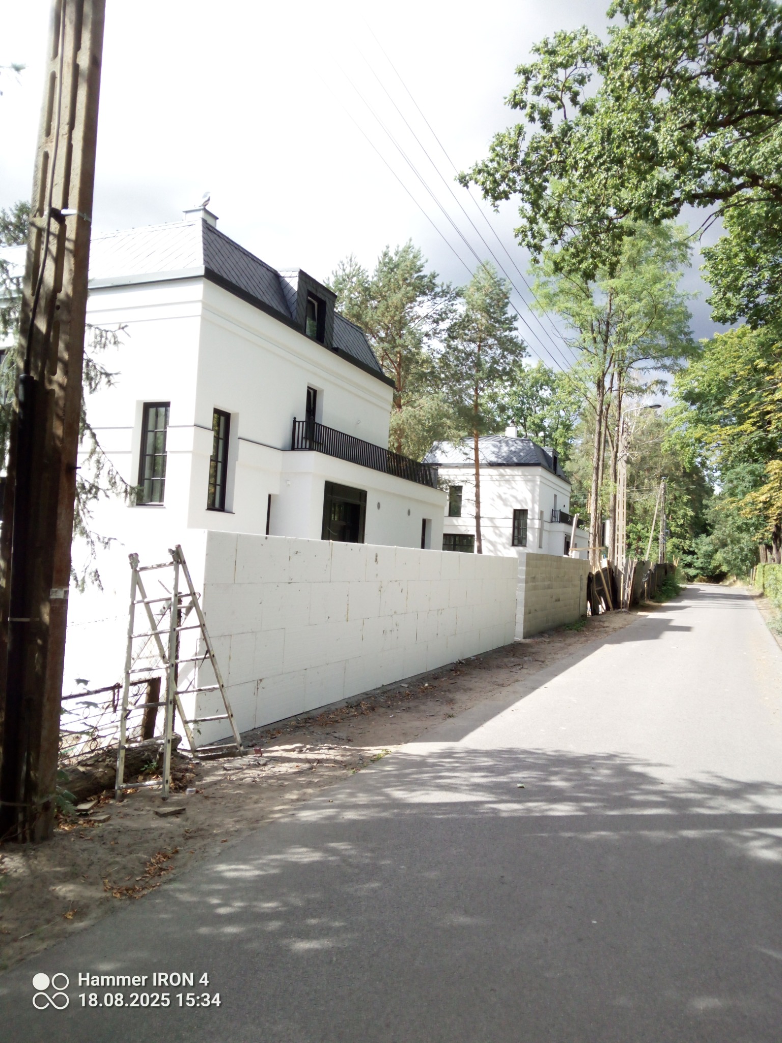 Nowoczesny biały dom z czarnym dachem, widoczny z ulicy za murem z białych bloczków. Drabina oparta o mur. Otoczenie zielone, drzewa. Słoneczny dzień.