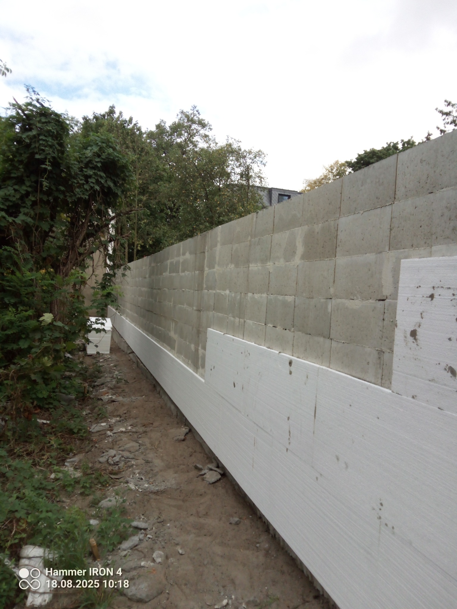 Ściana z szarych bloczków betonowych z częściowo zamontowaną białą izolacją styropianową, widok z dołu, obok ziemia i zieleń.