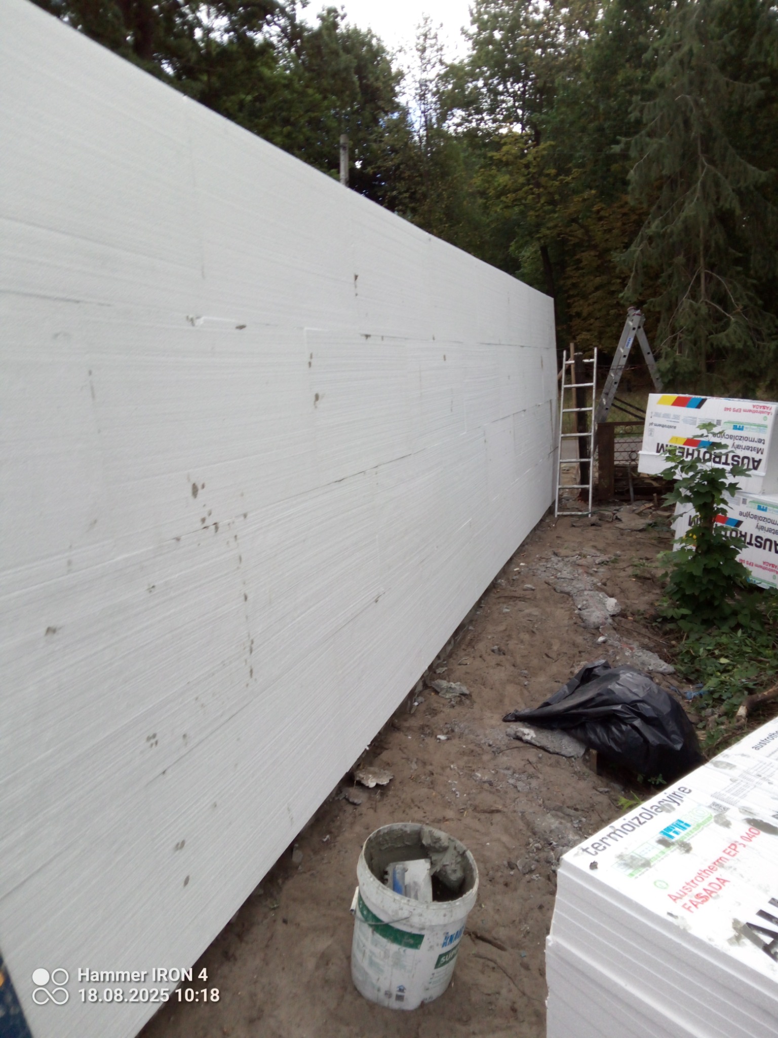 Ocieplona ściana styropianem, widoczna drabina, zaprawa w wiadrze i paczki styropianu. Prace ociepleniowe w toku, tło z drzewami.