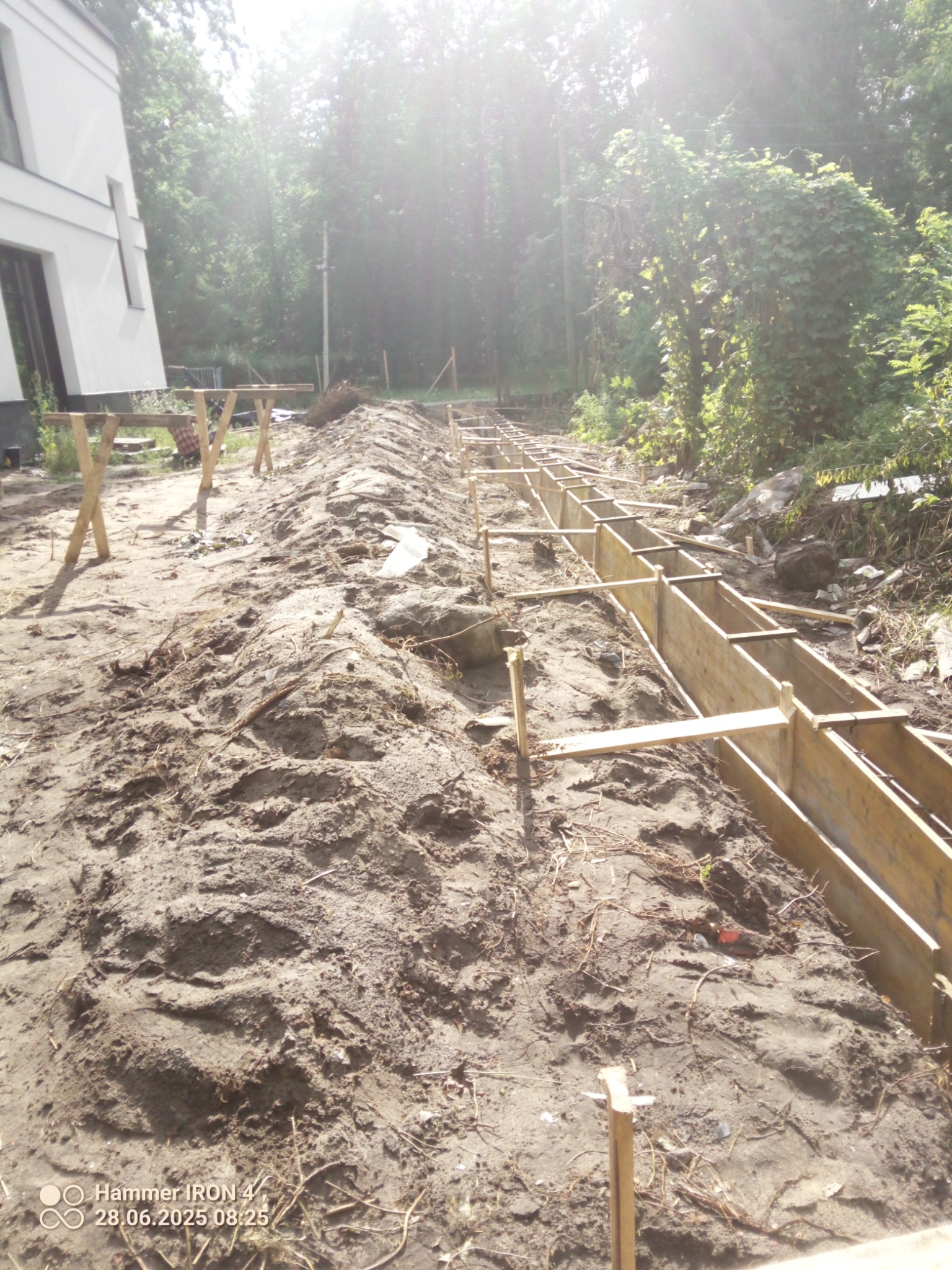 Przygotowanie terenu pod fundament: drewniane szalunki w wykopie, piasek, w tle zieleń i budynek. Widok z góry, jasne światło.