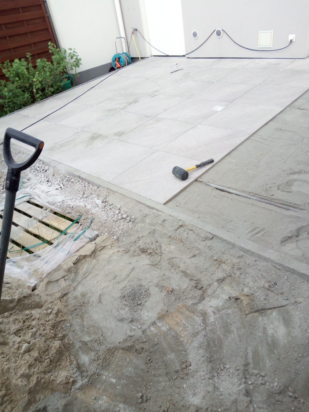 Układanie jasnoszarej kostki brukowej na zewnątrz budynku. Widoczny gumowy młotek, łopata i piasek. Częściowo ułożona nawierzchnia. Prace brukarskie w toku.
