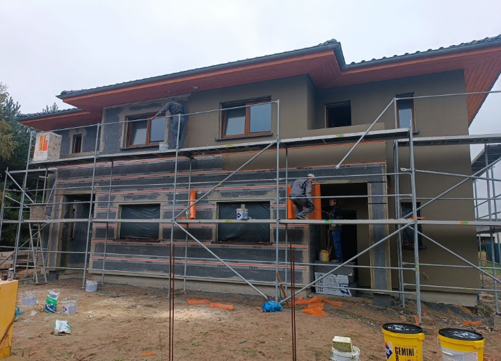 Dom w trakcie ocieplania, widoczne rusztowania i pracownicy. Część elewacji otynkowana na szaro, okna z brązowymi ramami. Na ziemi wiadra z farbą.