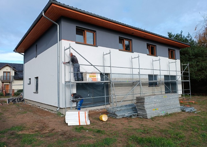 Dwóch pracowników na rusztowaniu ociepla białą elewację domu z szarą górną częścią. Widoczny styropian, narzędzia i elementy rusztowania. Dom z brązowymi oknami.