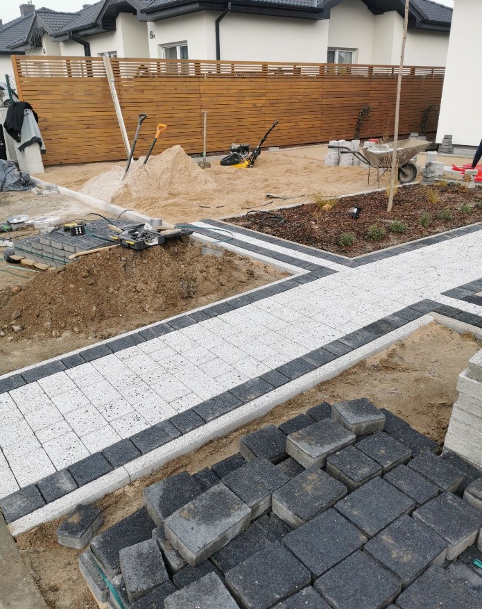 Etap układania jasnej kostki brukowej z ciemną obwódką na posesji. Widoczny piasek, narzędzia, częściowo ułożony chodnik i drewniane ogrodzenie w tle.