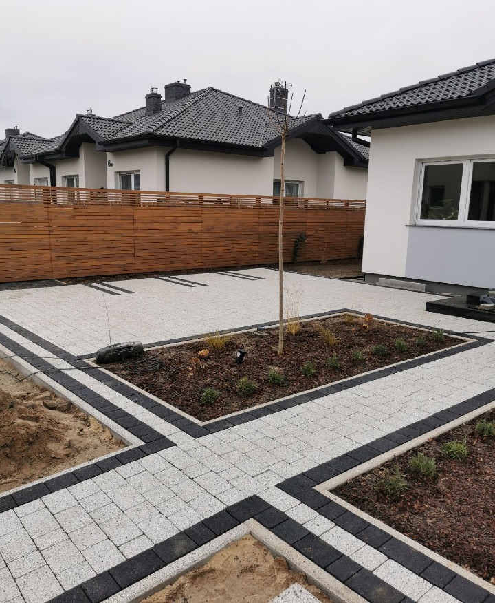 Nowoczesny podjazd z jasnej kostki brukowej z czarnymi obrzeżami, rabata z roślinami pośrodku, drewniane ogrodzenie w tle, domy jednorodzinne.