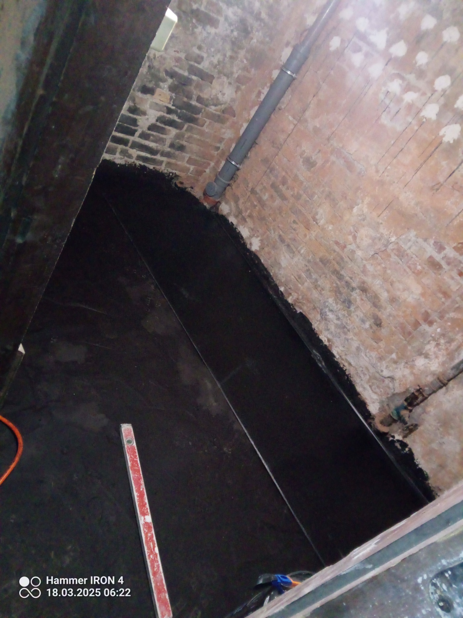 Zabezpieczenie fundamentów czarną folią hydroizolacyjną przy ceglanej ścianie. Widoczny poziomica i fragment węża. Narożne ujęcie z góry.