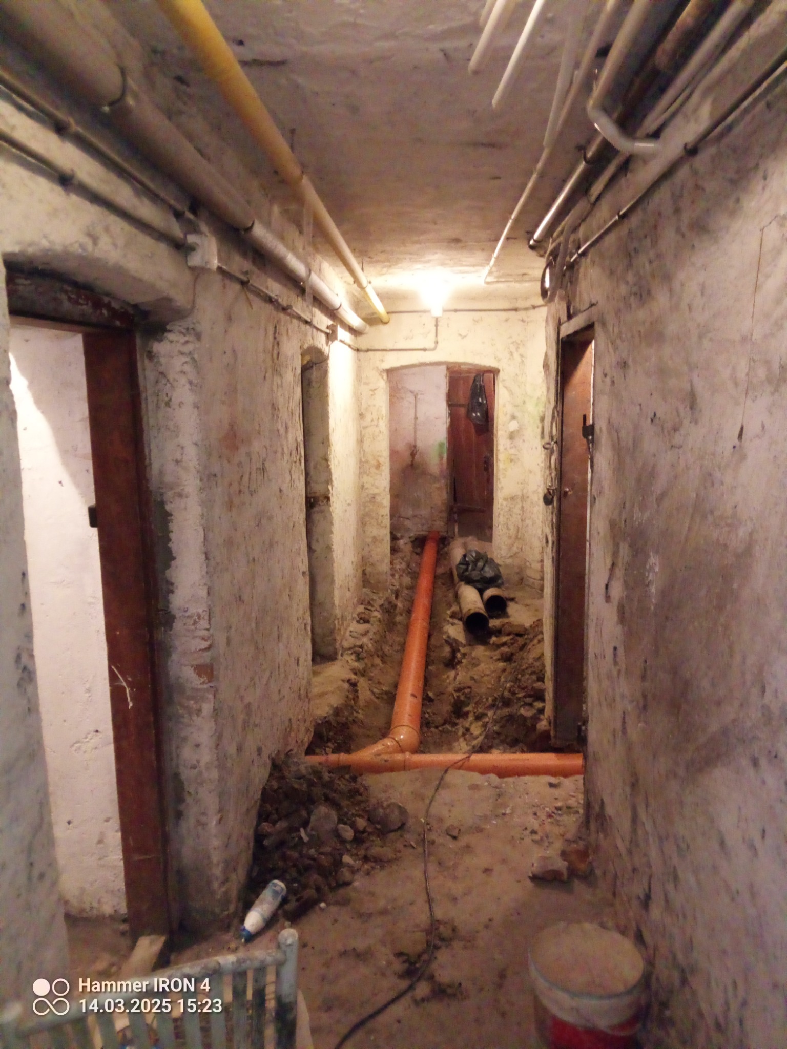 Pomarańczowe rury kanalizacyjne ułożone w wykopie w piwnicy, widoczne ściany i rury pod sufitem. Etap prac instalacyjnych w surowym wnętrzu.