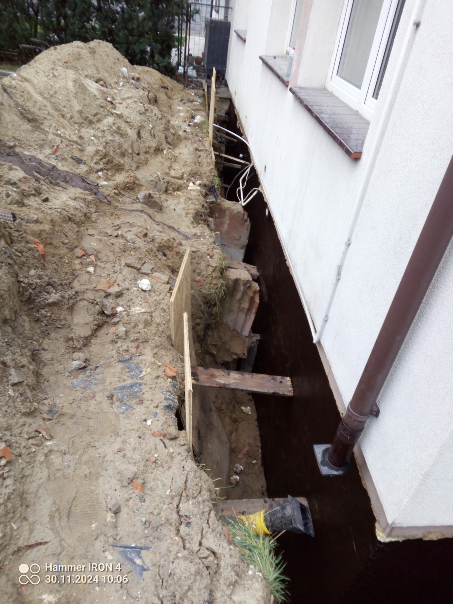 Odkopana ściana fundamentowa budynku z izolacją bitumiczną, zabezpieczona deskami. Widoczny rów z ziemią i wystające elementy instalacji. Prace ziemne.