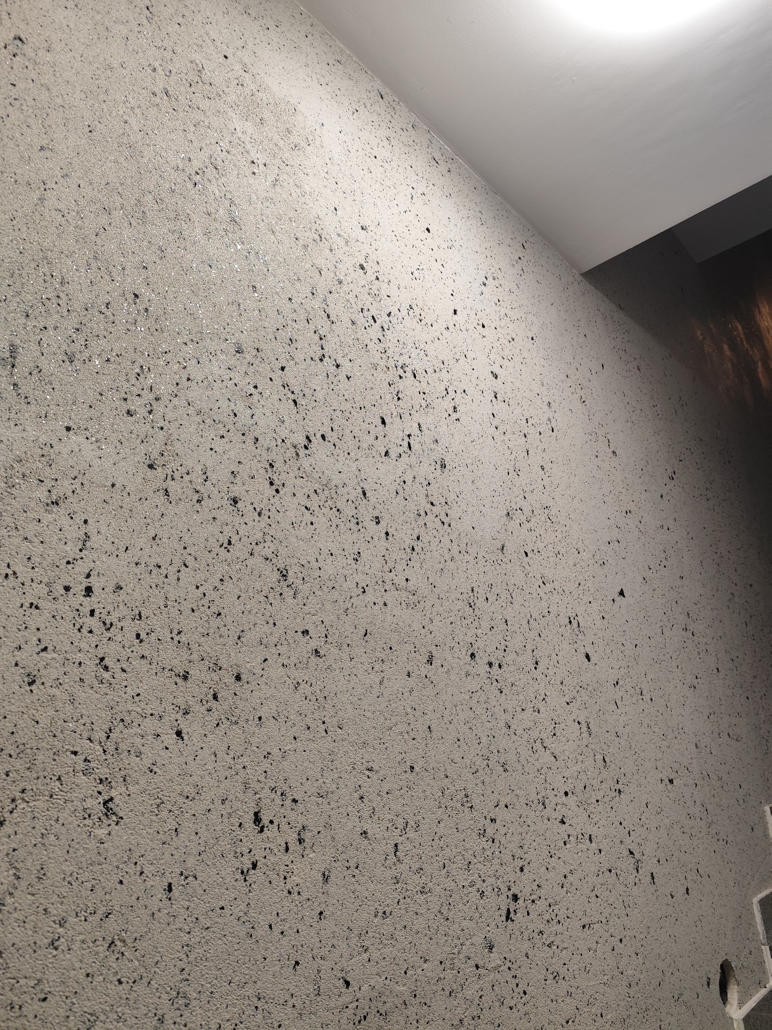 Szara ściana z fakturą i czarnymi plamkami, oświetlona od góry. Widoczny fragment sufitu. Tekstura ściany jest nierówna, z widocznymi drobnymi wgłębieniami.