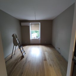 KJM-INSTAL - Pokój po remoncie z drewnianą podłogą, świeżo pomalowanymi ścianami w odcieniach beżu i szarości, widocznym klimatyzatorem oraz drabina malarska z pozostawioną szmatką, sugerująca prace wykończeniowe.