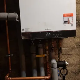 Nowoczesny, biały piec grzewczy Viessmann zamontowany na szarej ścianie, z widocznymi miedzianymi rurami, zaworami i izolacją termiczną, wraz z naklejką 'Connect me!' promującą aplikację mobilną.