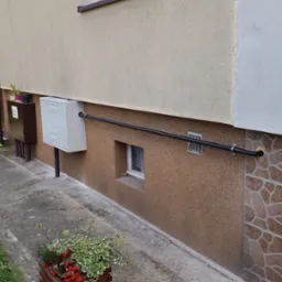 Zewnętrzna instalacja gazowa z czarną rurą przytwierdzoną do ściany budynku, z widoczną skrzynką gazową i fragmentem elewacji z oknem i kwiatami w donicy na pierwszym planie.