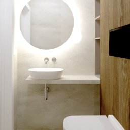 Agata Ambroziak - Banicka CREATIONS - Minimalistyczna łazienka z okrągłym lustrem podświetlanym LED, wiszącą umywalką i toaletą podwieszaną, wykończona drewnem i jasnym gresem.