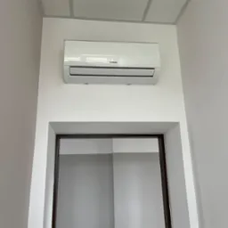 Biały klimatyzator marki Kaisai zamontowany na ścianie nad drzwiami w jasnym pomieszczeniu z kasetonowym sufitem. Widok od dołu.