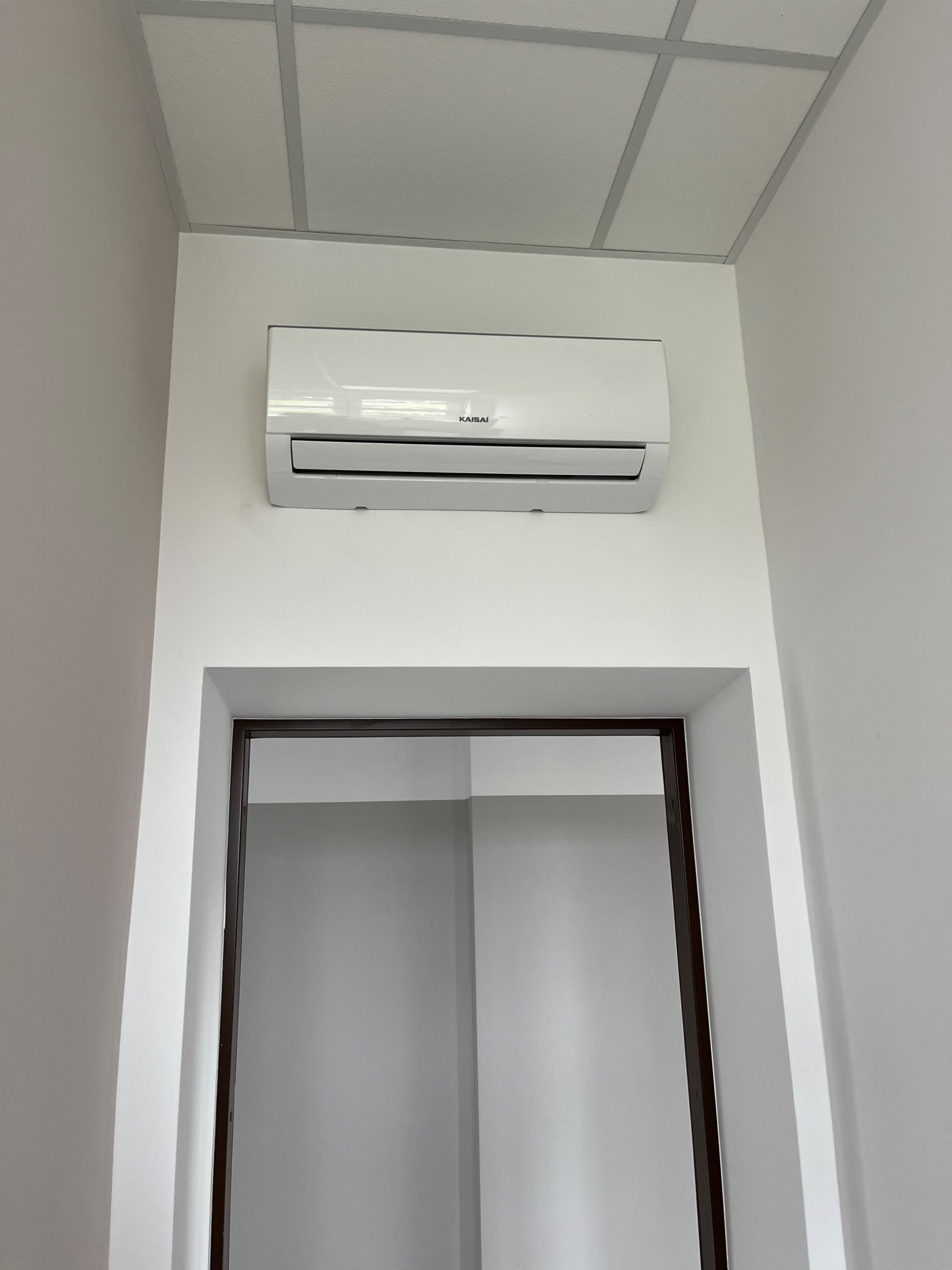 Biały klimatyzator marki Kaisai zamontowany na ścianie nad drzwiami w jasnym pomieszczeniu z kasetonowym sufitem. Widok od dołu.