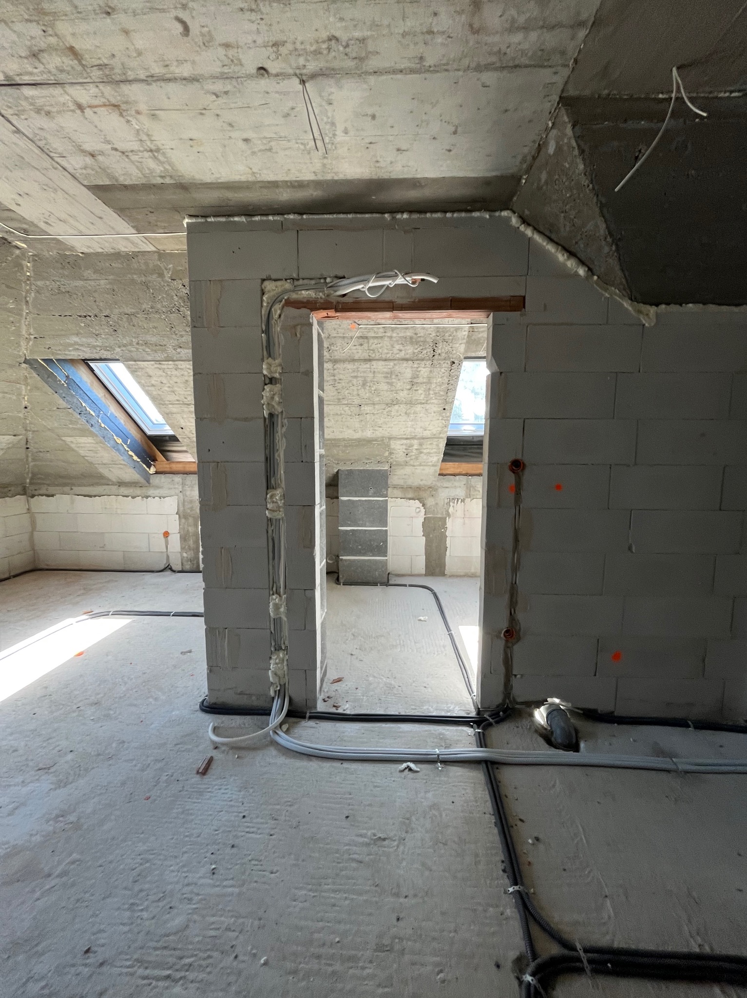 Wnętrze w trakcie prac elektrycznych: przewody ułożone wzdłuż ścian z bloczków, widoczne puszki instalacyjne i okno dachowe, surowy betonowy strop.