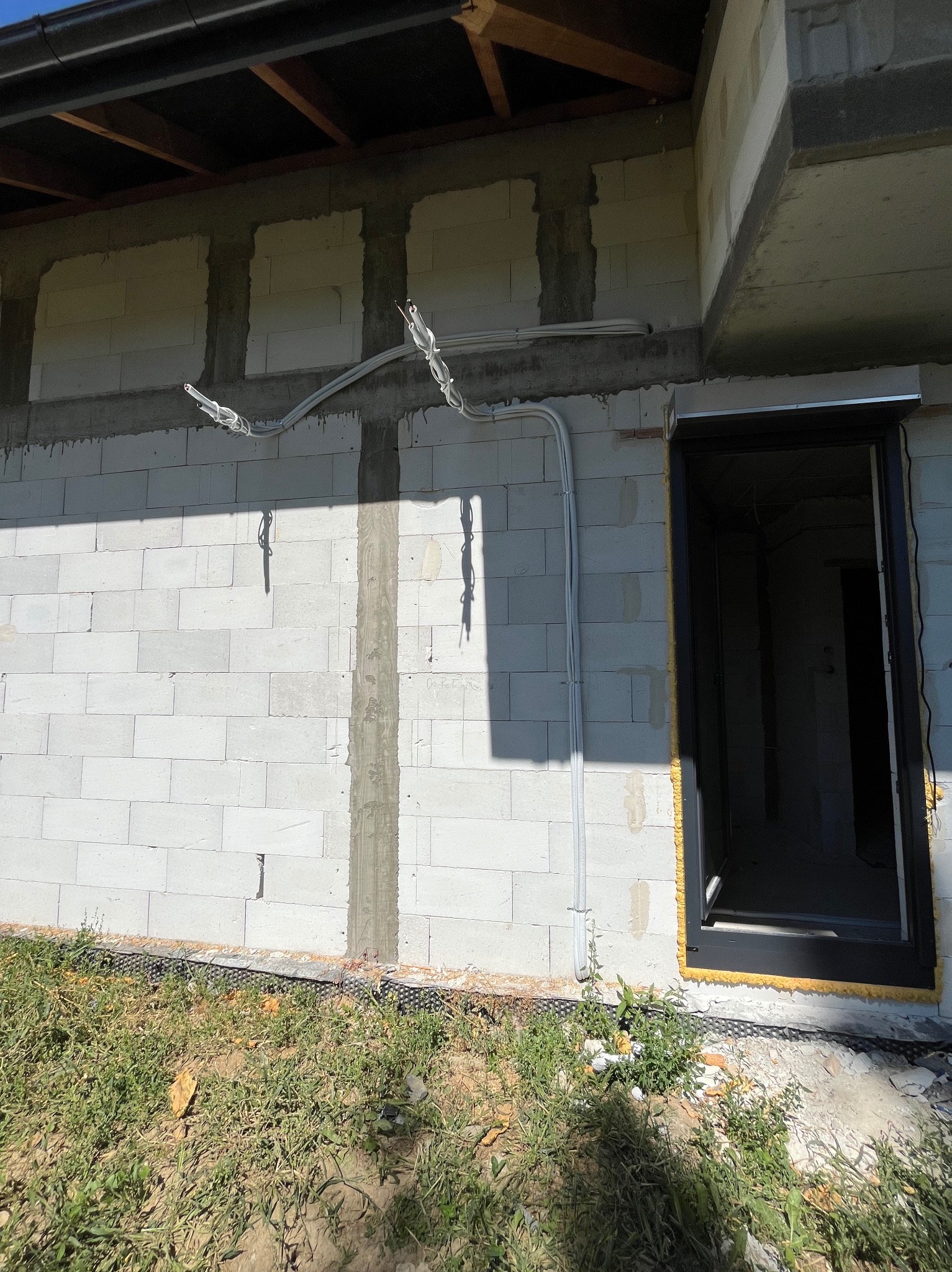 Biały budynek w trakcie budowy z widocznymi przewodami elektrycznymi na ścianie i czarną ramą okienną z żółtą pianką montażową.