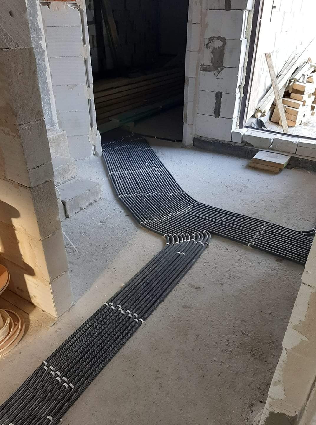 Rzędy szarych rur karbowanych z białymi złączkami ułożone na surowym betonowym podłożu wewnątrz budynku w trakcie budowy, widoczne elementy konstrukcyjne z betonu komórkowego.