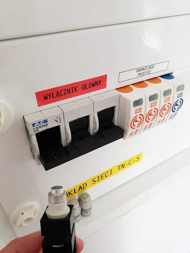 Otwarta skrzynka bezpiecznikowa z wyłącznikiem głównym Eaton, ogranicznikiem przepięć i bezpiecznikami. W dłoni bezpiecznik. Żółta naklejka z napisem 'Układ sieci TN-C-S'.