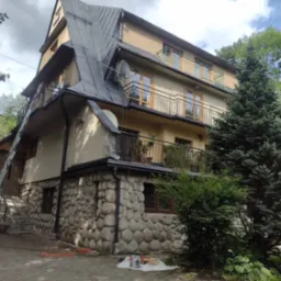 Wielopiętrowy dom z ciemnym, skośnym dachem, widoczne narzędzia i drabina sugerujące prace na wysokości, elewacja częściowo kamienna, otoczony zielenią.