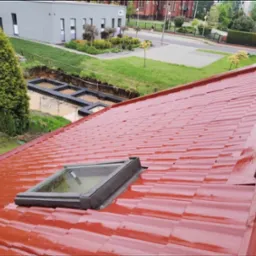 Czerwony, błyszczący dach z dachówki trapezowej po malowaniu, z widocznym oknem dachowym i fragmentem rynny, w tle budynek i wykopy fundamentowe.