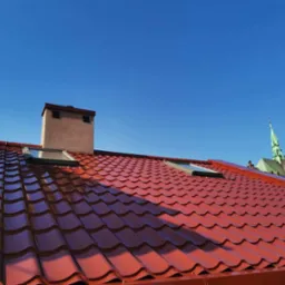 Czerwony dach z blachodachówki z oknami dachowymi i kominem na tle błękitnego nieba, w tle widoczna wieża kościoła.