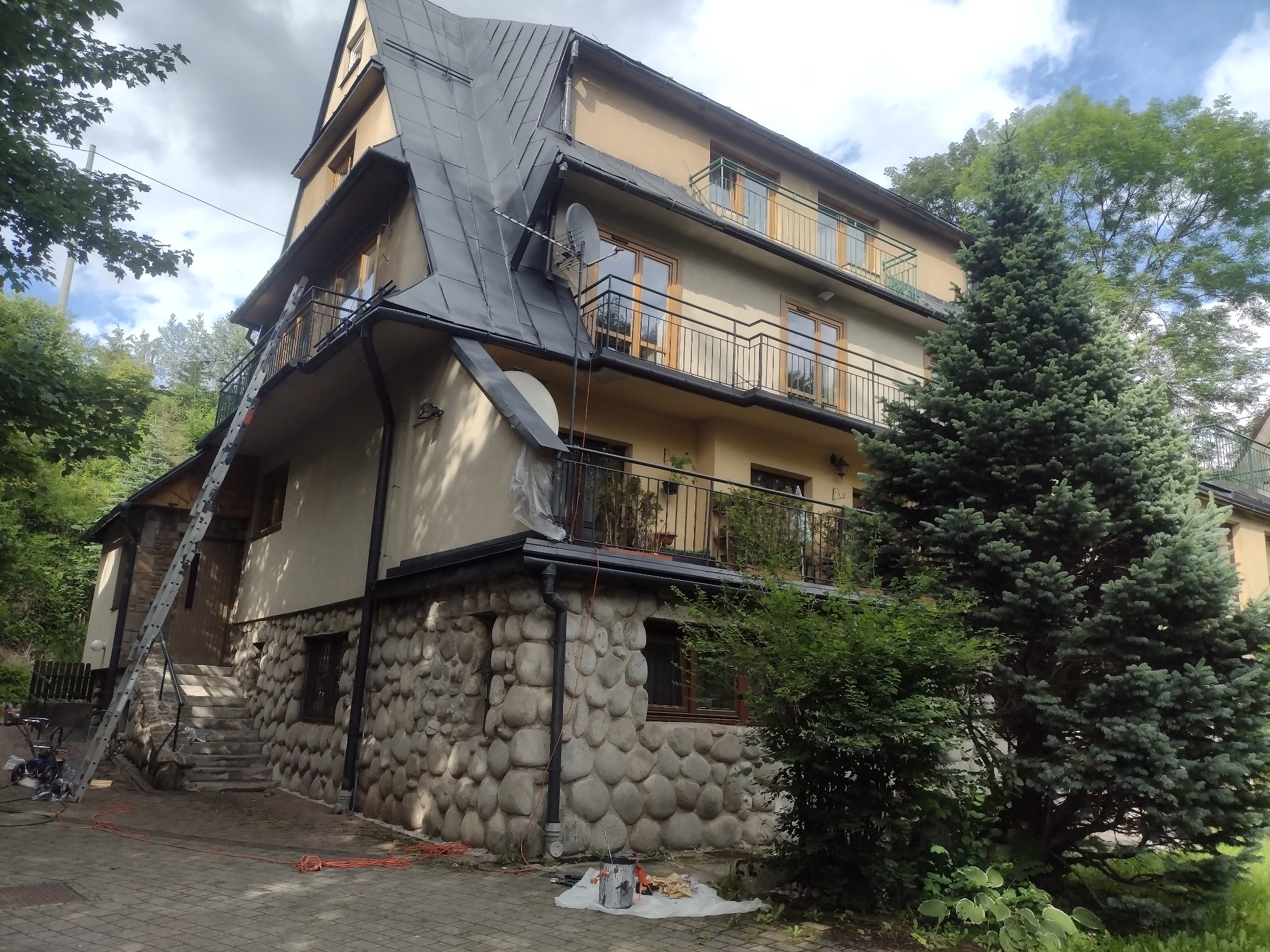 Wielopiętrowy dom z ciemnym, skośnym dachem, widoczne narzędzia i drabina sugerujące prace na wysokości, elewacja częściowo kamienna, otoczony zielenią.