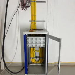 Szafa rack z uporządkowanymi, żółtymi kablami wewnątrz, widoczne narzędzia na górze szafy, instalacja kablowa na ścianie. Otwarta przeszklona obudowa.