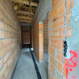 Michalski-Elektro Zbigniew Michalski - Wnętrze w trakcie budowy: widok korytarza z instalacją elektryczną w bruzdach ścian z cegły, widoczne kable w peszlach i puszki elektryczne, surowy beton i drewniana konstrukcja dachu.